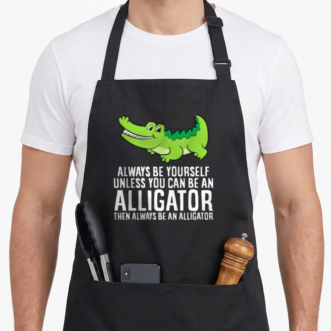 Crocodile unless apron - Green cartoon alligator above white text ALWAYS BE YOURSELF UNLESS YOU CAN BE AN ALLIGATOR — shown on a black cotton canvas apron against a white background