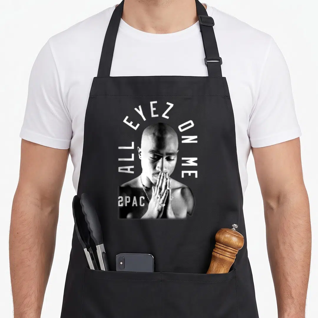 Against tupac apron - Grayscale man praying with ALL EYEZ ON ME text and 2PAC logo — shown on a black cotton canvas apron against a white background