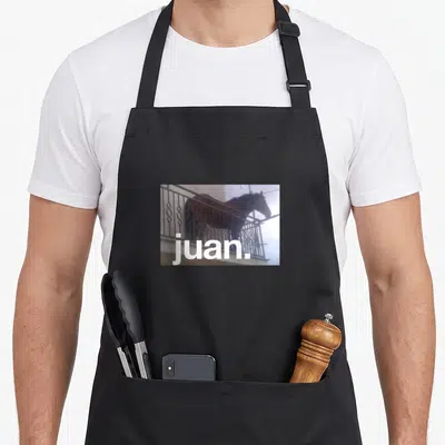 Juan horse apron - Brown horse standing behind grey metal fence with white text juan — shown on a black cotton canvas apron against a white background