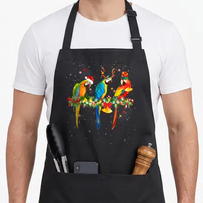 Colorful christmas elf apron - Three parrots in red green blue yellow on Christmas garland with bells and candy canes — shown on a black cotton canvas apron against a white background