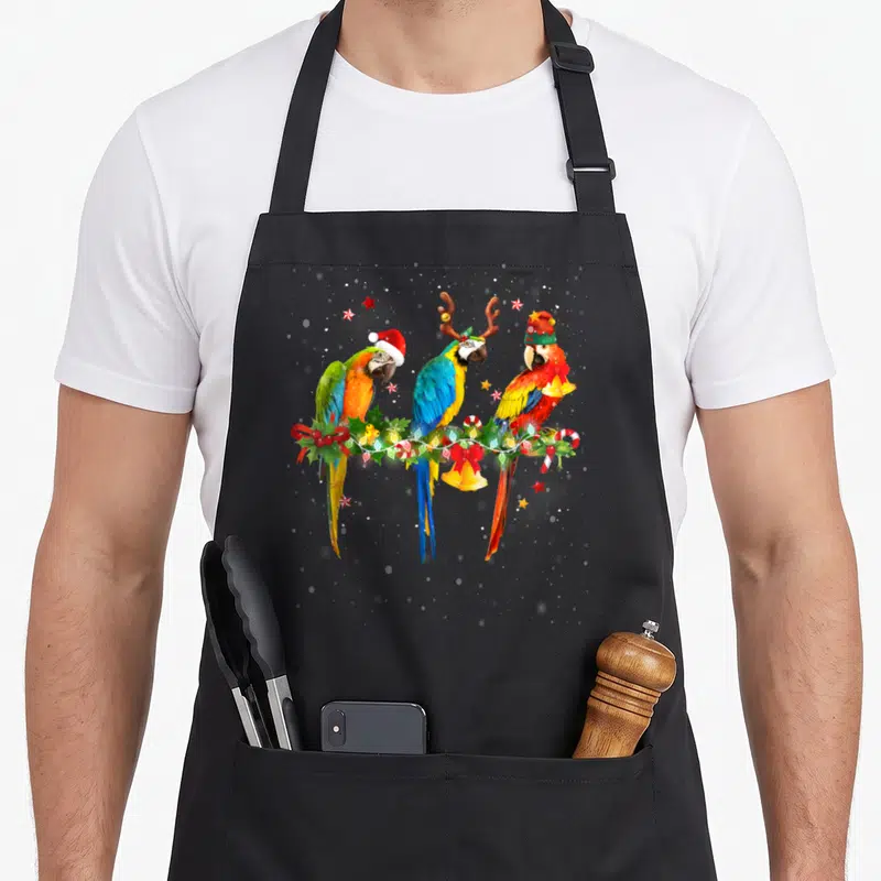 Colorful christmas elf apron - Three parrots in red green blue yellow on Christmas garland with bells and candy canes — shown on a black cotton canvas apron against a white background
