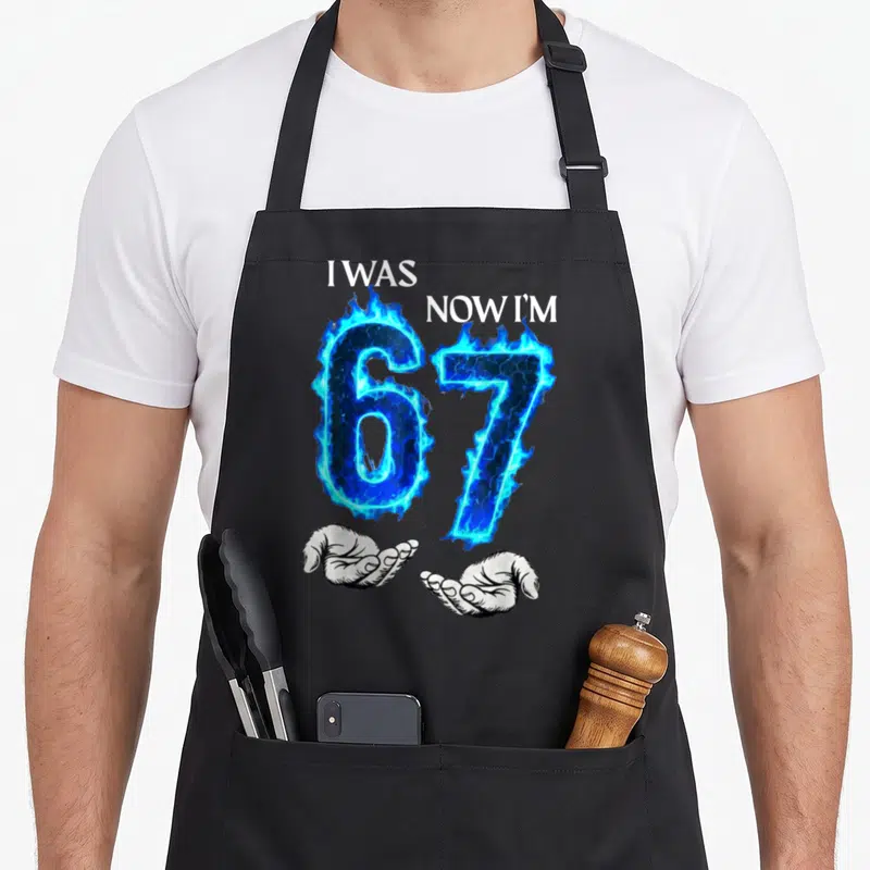 Cute general birthday apron for grilling - Blue flaming 67 held by two white hands with I WAS NOW I'M text — shown on a black cotton canvas apron against a white background
