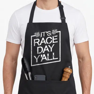 Race track apron - White stacked text IT'S RACE DAY Y'ALL in distressed frame with two checkered flags — shown on a black cotton canvas apron against a white background