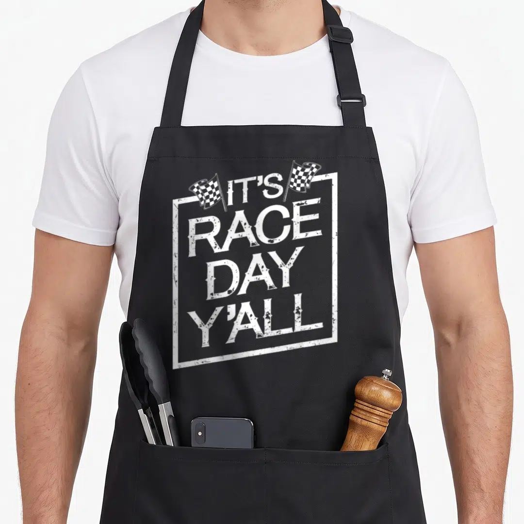 Race track apron - White stacked text IT'S RACE DAY Y'ALL in distressed frame with two checkered flags — shown on a black cotton canvas apron against a white background