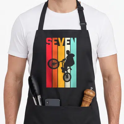 Bmx birthday apron - SEVEN text over red orange yellow cream blue stripes with grey bike wheelie silhouette — shown on a black cotton canvas apron against a white background