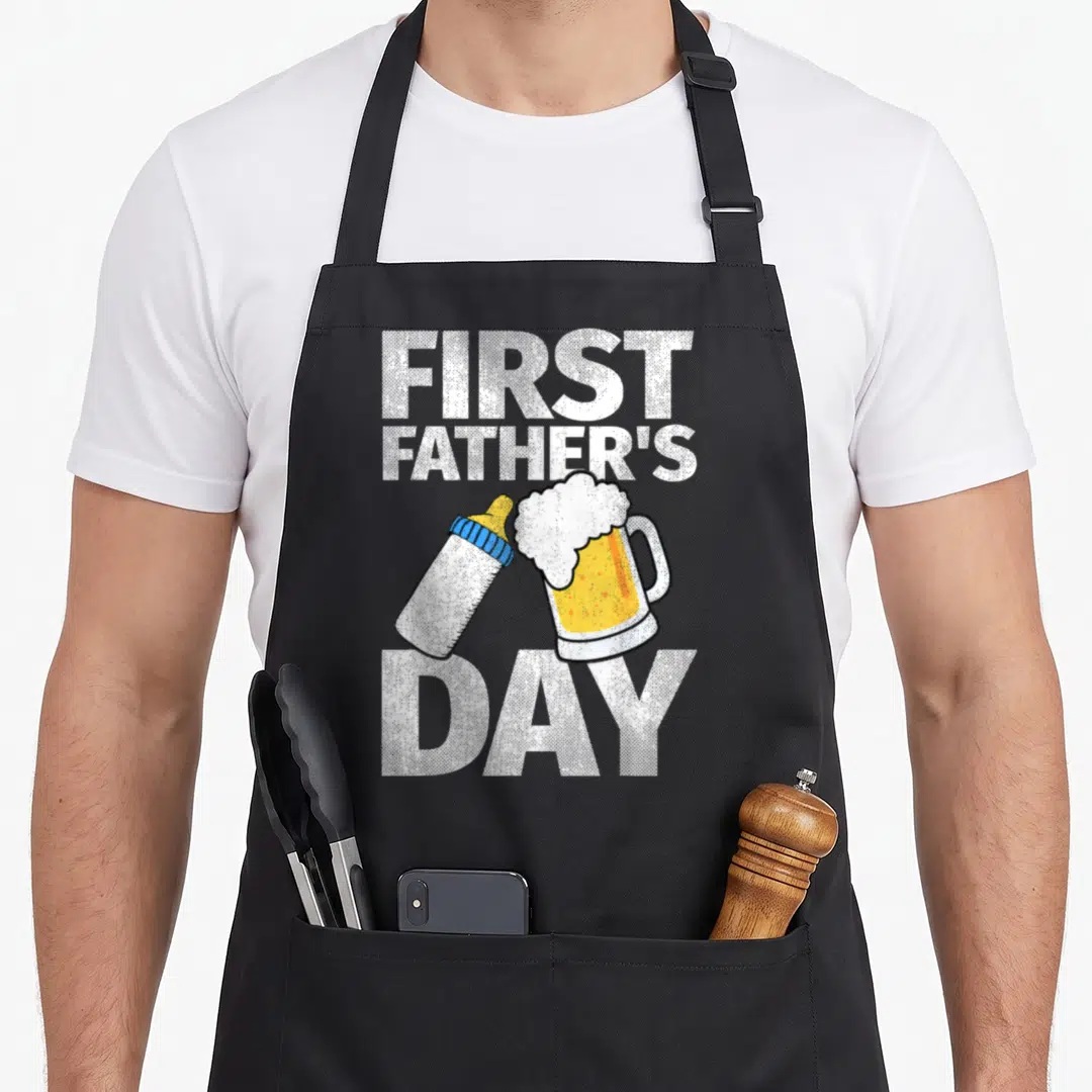 Beer father day apron - First Father's Day text with clinking baby bottle and beer mug in white yellow blue — shown on a black cotton canvas apron against a white background