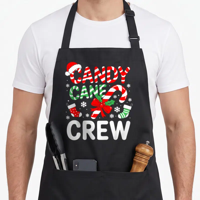 Pajamas candy cane apron - CANDY CANE CREW text with Santa hat, red bow, two stockings, snowflakes in red green white — shown on a black cotton canvas apron against a white background
