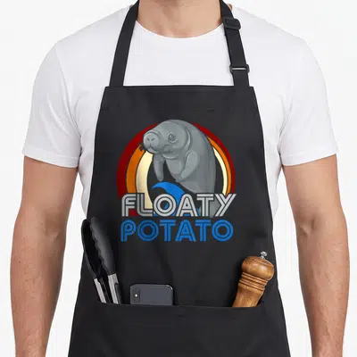 Floaty manatee apron - Grey cartoon manatee over red orange circle with blue wave and stacked text — shown on a black cotton canvas apron against a white background