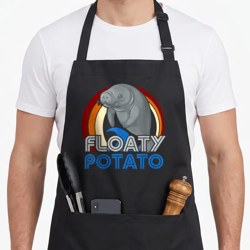 Floaty manatee apron - Grey cartoon manatee over red orange circle with blue wave and stacked text — shown on a black cotton canvas apron against a white background