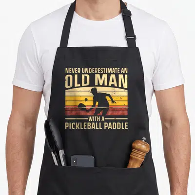 Pickleball grandpa apron - Retro sunset stripes with grey pickleball player silhouette and bold beige text declaring — shown on a black cotton canvas apron against a white background