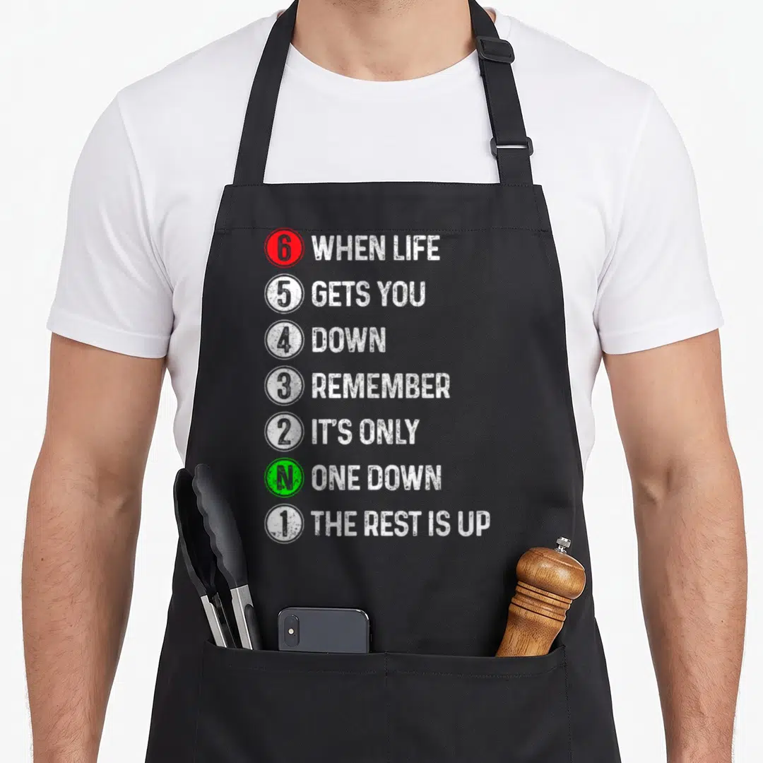 Gear motorcycle apron - Stacked white text with red, green, and white numbered circles reading 6 when life gets you down — shown on a black cotton canvas apron against a white background