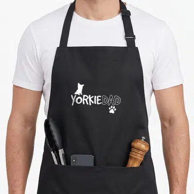 Owner yorkie apron - White YORKIEDAD text with sitting dog silhouette and paw print below — shown on a black cotton canvas apron against a white background