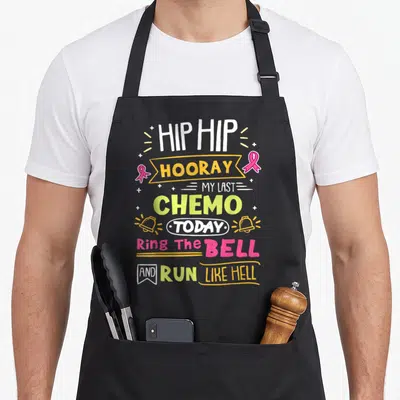 Chemo ring apron - Stacked text Hip Hooray My Last Chemo Today with pink ribbons and bells in white yellow pink lime — shown on a black cotton canvas apron against a white background