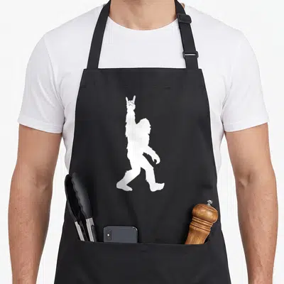 Sasquatch believer bigfoot apron - White silhouette of walking Bigfoot holding up a rock-on hand sign — shown on a black cotton canvas apron against a white background