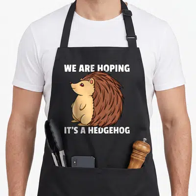 Pregnant hedgehog mom apron - Cartoon hedgehog with white text we are hoping its a hedgehog — shown on a black cotton canvas apron against a white background