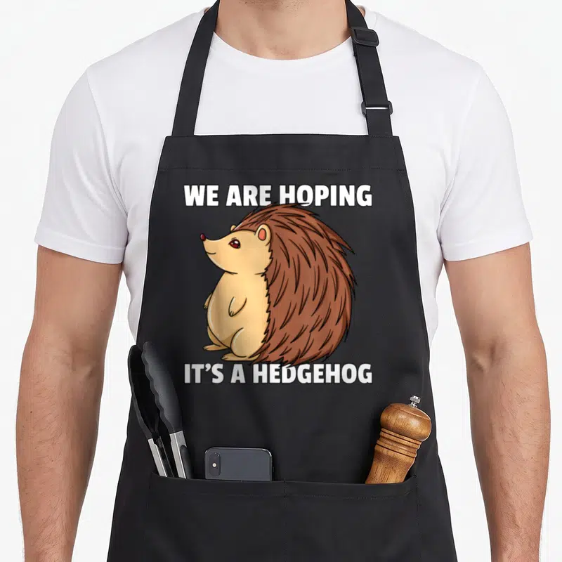Pregnant hedgehog mom apron - Cartoon hedgehog with white text we are hoping its a hedgehog — shown on a black cotton canvas apron against a white background