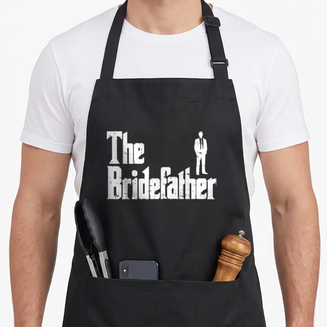 Rehearsal bride apron - White distressed text reading The Bridefather with man silhouette standing on letters — shown on a black cotton canvas apron against a white background