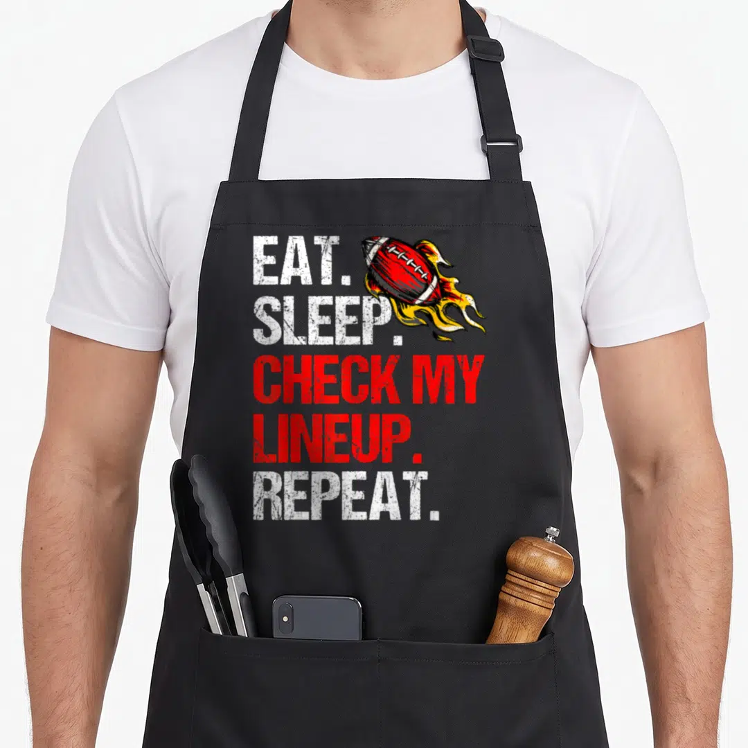 Sleep football apron - Stacked eat sleep check lineup repeat text next to a red flaming football — shown on a black cotton canvas apron against a white background