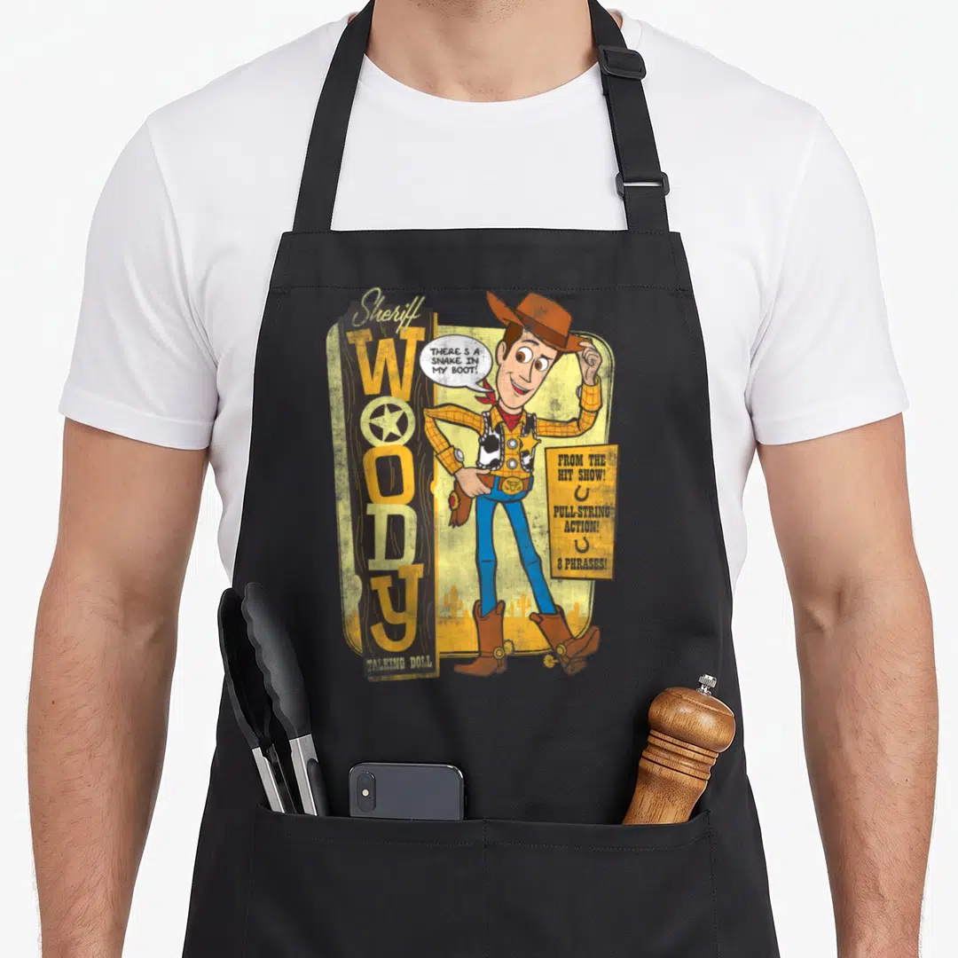 Sheriff doll apron - Cartoon Woody cowboy with snake in boot speech bubble and pull string action sign — shown on a black cotton canvas apron against a white background