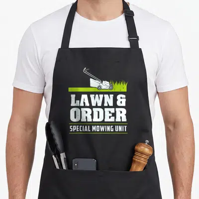 Landscaper mower lover apron - White lawn mower on green grass strip above stacked LAWN & ORDER text — shown on a black cotton canvas apron against a white background