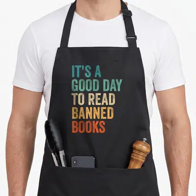 Read bibliophile apron - Stacked colorful text reading IT'S A GOOD DAY TO READ BANNED BOOKS in distressed block letters — shown on a black cotton canvas apron against a white background