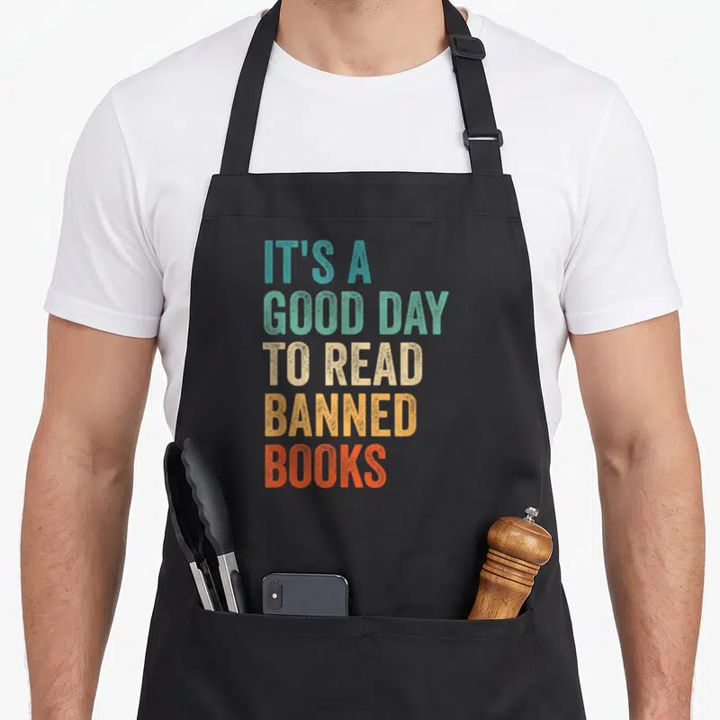 Read bibliophile apron - Stacked colorful text reading IT'S A GOOD DAY TO READ BANNED BOOKS in distressed block letters — shown on a black cotton canvas apron against a white background