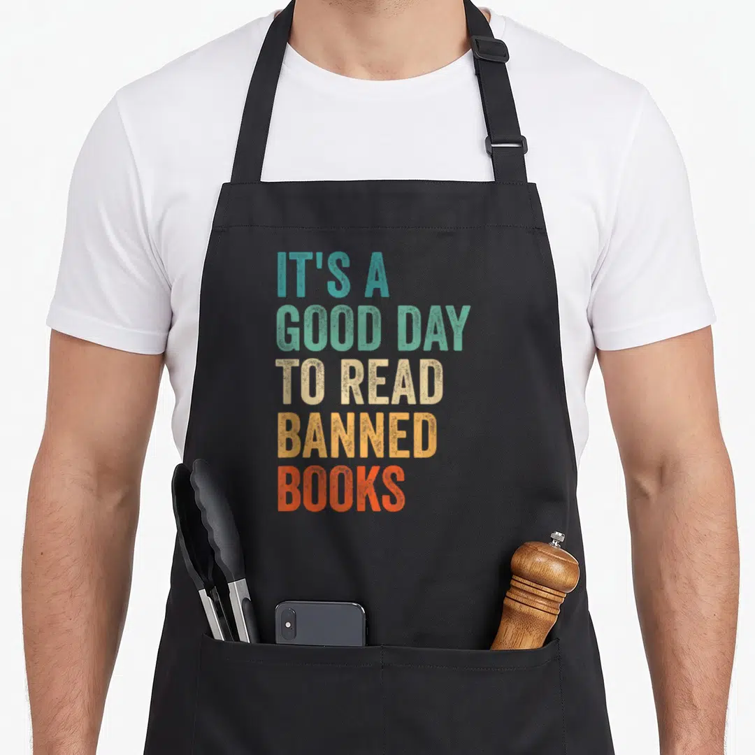 Read bibliophile apron - Stacked colorful text reading IT'S A GOOD DAY TO READ BANNED BOOKS in distressed block letters — shown on a black cotton canvas apron against a white background
