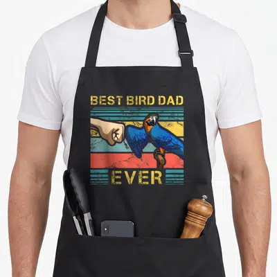 Enthusiast bird apron - Blue parrot fist bumping tan hand with Best Bird Dad Ever text on striped yellow blue red — shown on a black cotton canvas apron against a white background