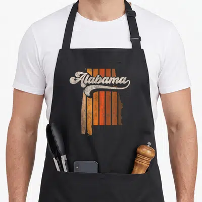 70s alabama apron - Retro script Alabama text over orange and brown vertical stripes inside a state map silhouette — shown on a black cotton canvas apron against a white background