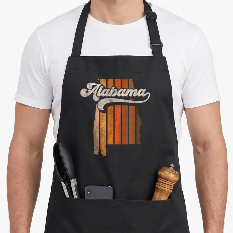 70s alabama apron - Retro script Alabama text over orange and brown vertical stripes inside a state map silhouette — shown on a black cotton canvas apron against a white background