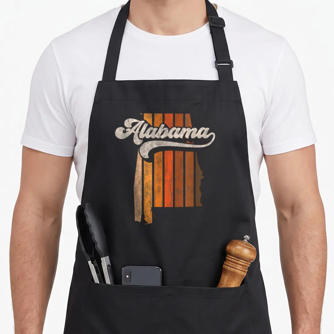 70s alabama apron - Retro script Alabama text over orange and brown vertical stripes inside a state map silhouette — shown on a black cotton canvas apron against a white background