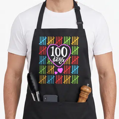 Counting teacher apron - 100 days text in pink heart surrounded by colored tally marks — shown on a black cotton canvas apron against a white background