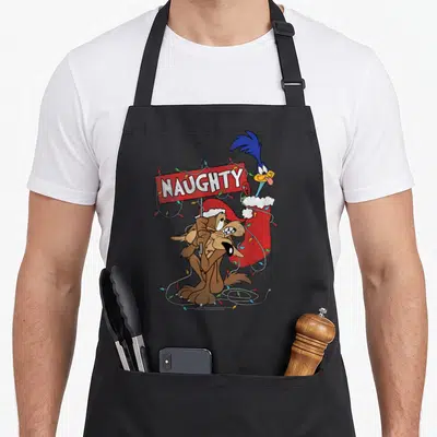 Road christmas apron - Wile E. Coyote holds red NAUGHTY sign with Road Runner in stocking and Christmas lights — shown on a black cotton canvas apron against a white background