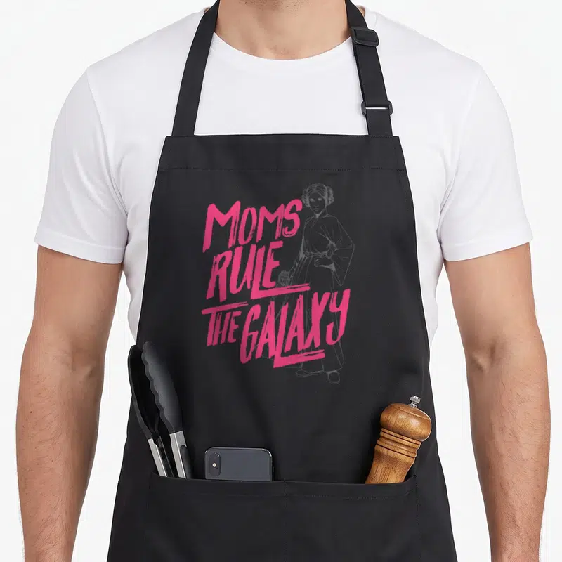 Leia mother's day aprons - Pink MOMS RULE THE GALAXY text beside grey line art of Princess Leia holding a blaster — shown on a black cotton canvas apron against a white background