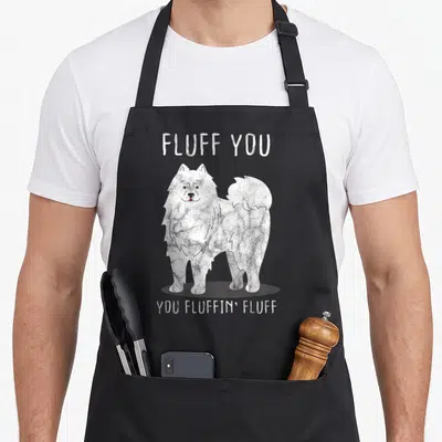 Fluffin dog apron - Cartoon white dog with text FLUFF YOU, YOU FLUFFIN' FLUFF on grey background — shown on a black cotton canvas apron against a white background