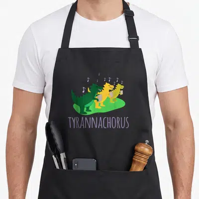 Chorus dinosaur apron - Three cartoon dinosaurs holding sheet music with purple TYRANNACHORUS text below — shown on a black cotton canvas apron against a white background