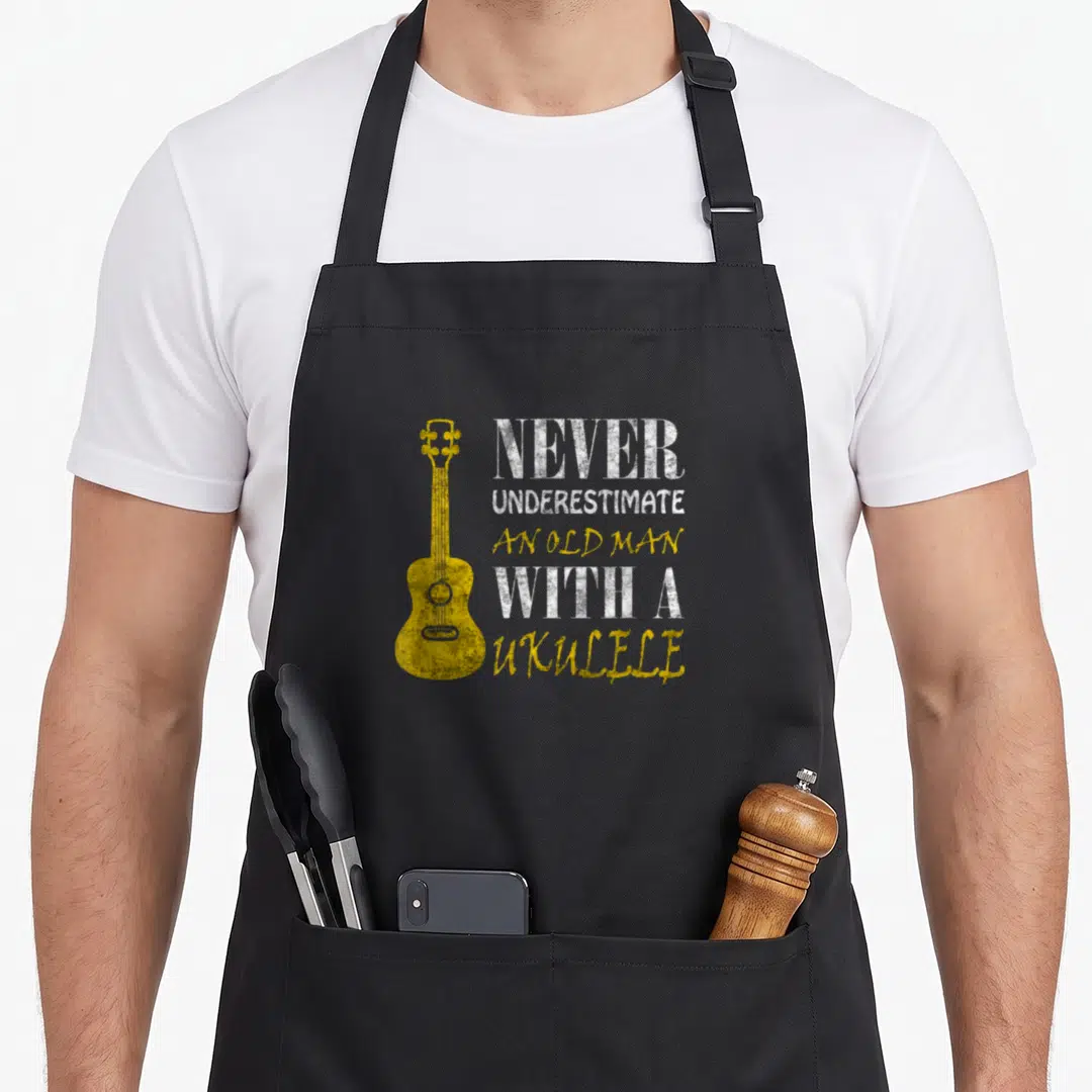 Underestimate ukulele apron - Yellow ukulele next to distressed white and yellow text reading Never Underestimate — shown on a black cotton canvas apron against a white background