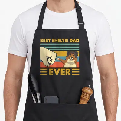 Sheltie pekingese apron - Retro fist bump between human hand and Sheltie dog on striped background with text — shown on a black cotton canvas apron against a white background