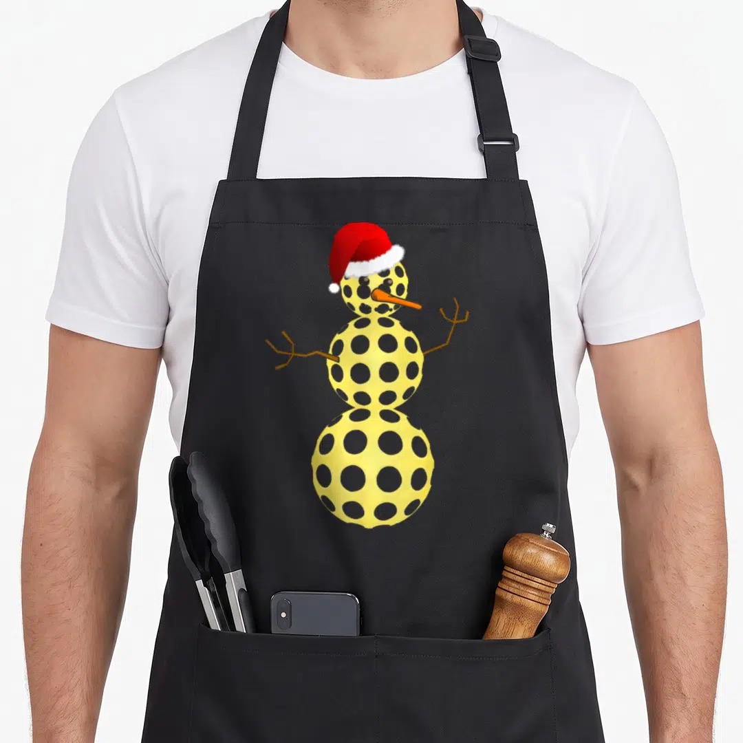 Pickleball snowman apron - Three tiered yellow snowman made of perforated balls with red santa hat and grey dots — shown on a black cotton canvas apron against a white background