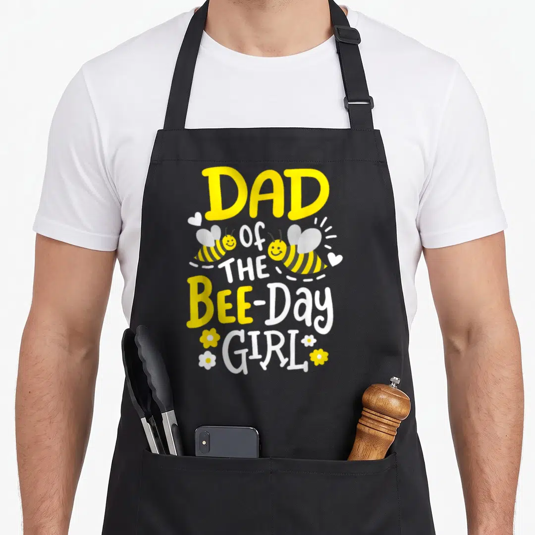 Birthday family bee aprons - DAD of THE BEE-Day GIRL text with two bees and scattered hearts and flowers in yellow — shown on a black cotton canvas apron against a white background