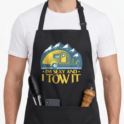 Camper motorhome owner apron - Retro yellow camper trailer with blue mountains and text I'm Sexy And I Tow It — shown on a black cotton canvas apron against a white background