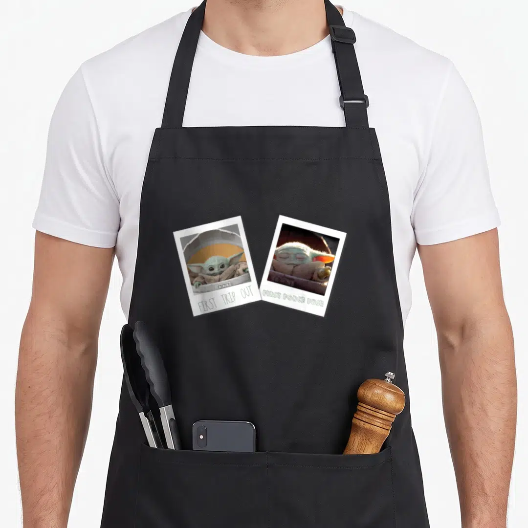 Funny memory apron - Two tilted polaroid photos of Grogu with captions FIRST TRIP OUT and FIRST FORCE PUSH — shown on a black cotton canvas apron against a white background