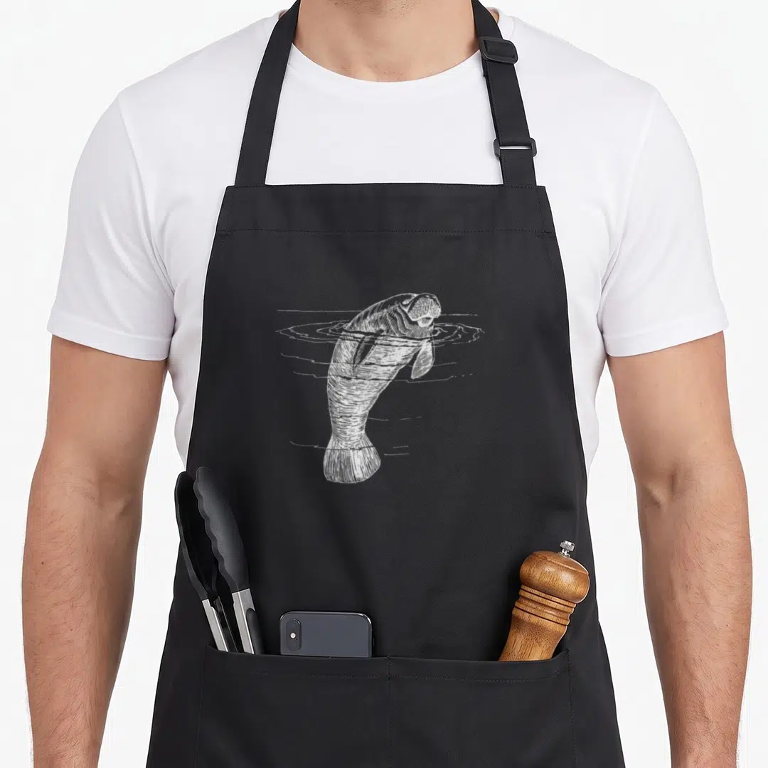 Illustration cow apron - White line drawing of a manatee swimming vertically with water ripples around its body — shown on a black cotton canvas apron against a white background