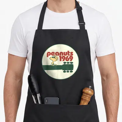 Woodstock machine apron - Snoopy character Woodstock sitting on dark green guitar neck inside cream circle with red text — shown on a black cotton canvas apron against a white background