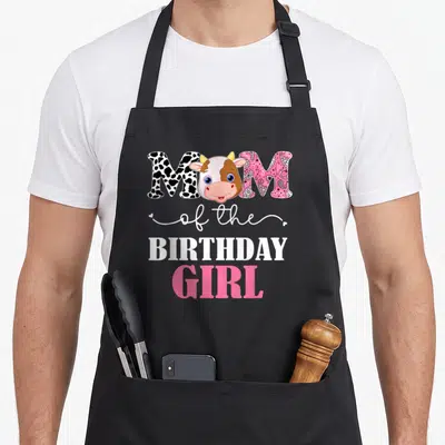 1st cow print apron - Cartoon cow head between spotted and paisley M letters with MOM of the BIRTHDAY GIRL text — shown on a black cotton canvas apron against a white background