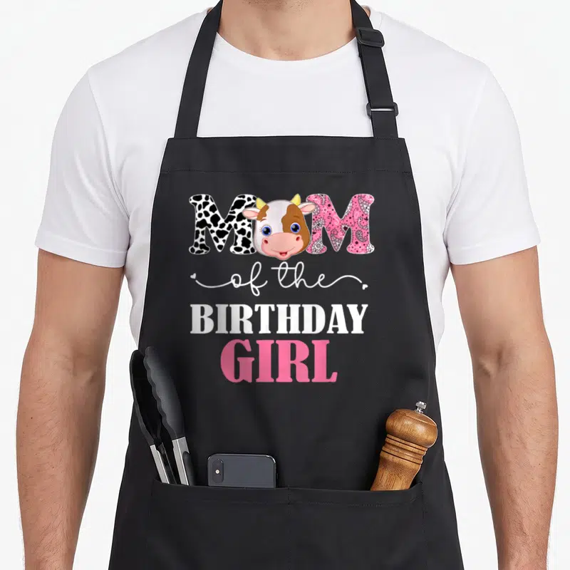 1st cow print apron - Cartoon cow head between spotted and paisley M letters with MOM of the BIRTHDAY GIRL text — shown on a black cotton canvas apron against a white background