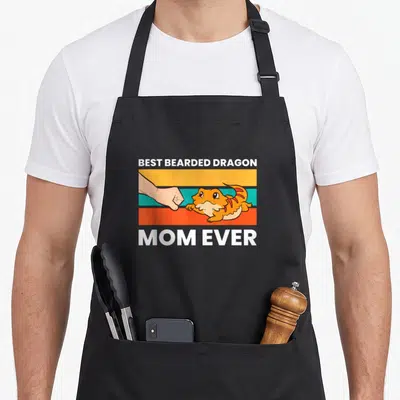Mum dragon apron - Cartoon orange bearded dragon fist bumping human hand on striped background — shown on a black cotton canvas apron against a white background