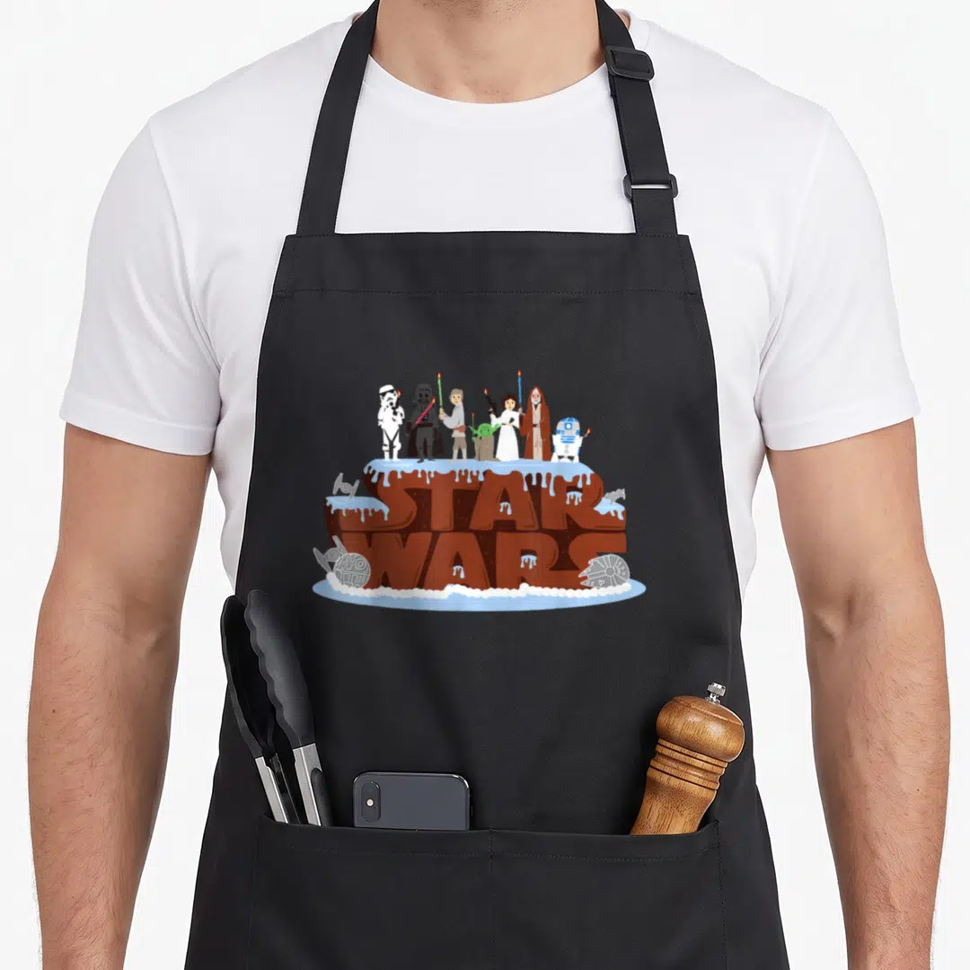 Character cake apron - Cartoon cake with STAR WARS text, characters holding candles, and spacecraft drawings on brown — shown on a black cotton canvas apron against a white background