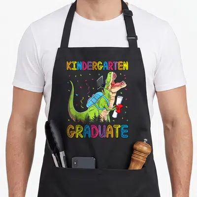 Graduate dinosaur apron - Green T-Rex in graduation cap holding diploma between arched colorful text — shown on a black cotton canvas apron against a white background