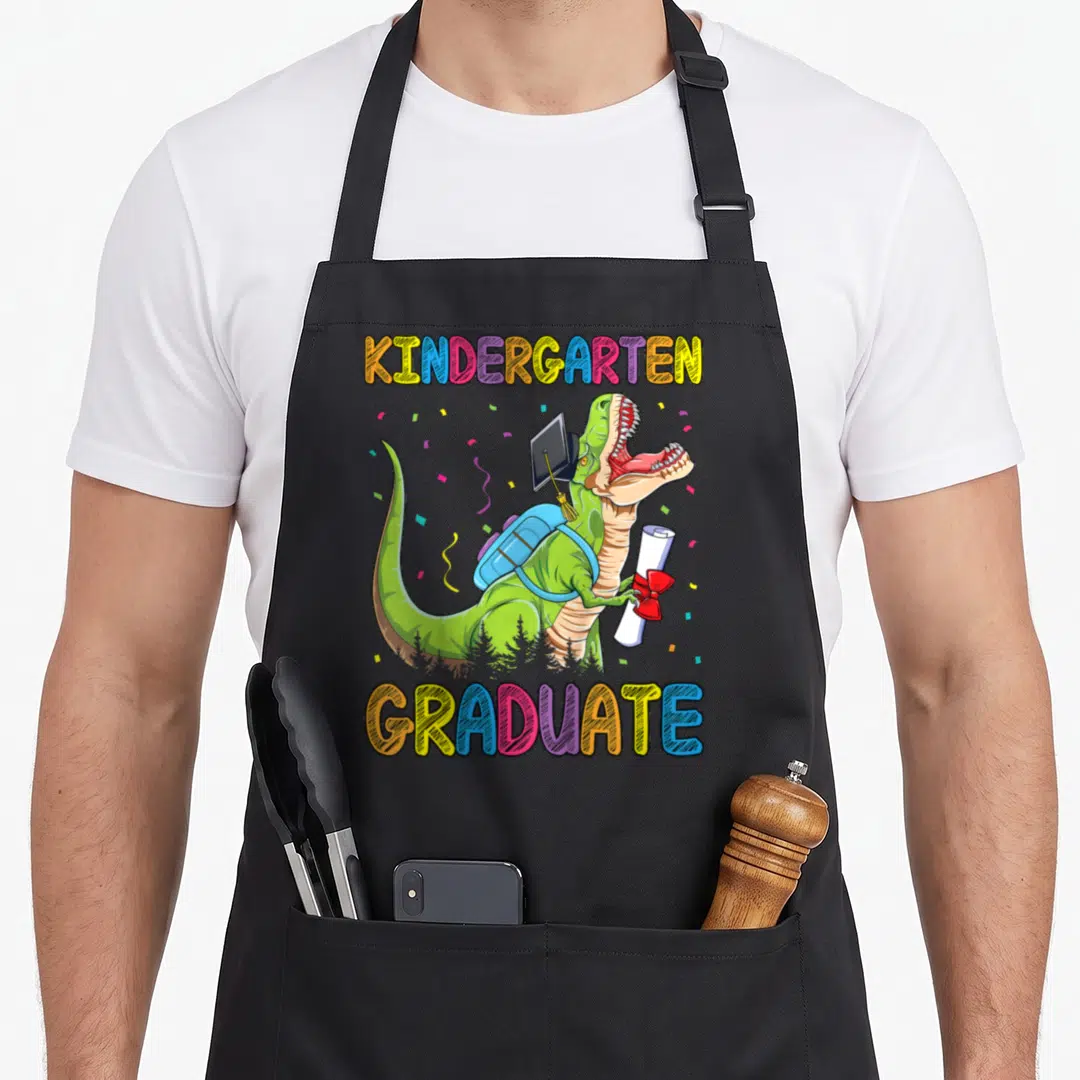Graduate dinosaur apron - Green T-Rex in graduation cap holding diploma between arched colorful text — shown on a black cotton canvas apron against a white background